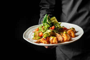 A white plate holds a gourmet dish featuring grilled shrimp, fresh salad greens, cherry tomatoes, and thinly sliced cucumber, garnished with red pepper flakes. The dark background