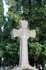 Close-up of a stone carving depicting the crucifixion of Jesus Christ with the inscription JHS. It features an elegant figure of the cross against a background of green cypress trees.