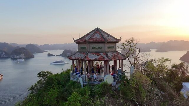 Scenic aerial footage of a traditional Vietnamese temple perched atop a mountain in Ha Long Bay. Tourists gather to admire the panoramic view of limestone islands and cruise boats at golden hour. A bl