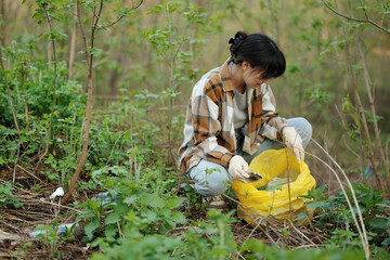 Young woman in casual clothes and gloves picking up trash in the forest using a yellow plastic bag....