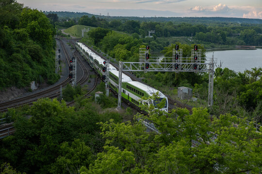 Aerial view of a train going through a lush green forest area by the lake in summer time at sunset hour. Commute, public transit, green transport, zero emission, travel, journey concept. 