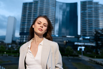 Fototapeta premium confident woman in beige blazer with modern cityscape background during evening light, urban style, business fashion, stylish professional young woman