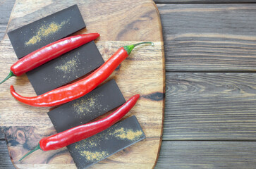 Bar of extra dark chocolate with three red chili peppers on a wooden cutting board on the table. Trend food concept. Selective focus. Horizontal orientation. Copy space. Top view