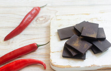 Pieces of extra dark chocolate with three red chili peppers on a white wooden cutting board on the table. Trend food concept. Selective focus. Horizontal orientation
