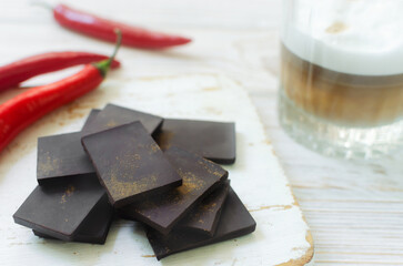 Pieces of extra dark chocolate with three red chili peppers on a white wooden cutting board with a glass of cappuccino on the table. Trend food concept. Selective focus. Horizontal orientation.