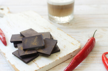 Pieces of extra dark chocolate with three red chili peppers on a white wooden cutting board with a glass of cappuccino on the table. Trend food concept. Selective focus. Horizontal orientation.