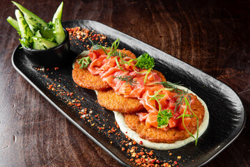 A gourmet dish of crispy potato pancakes topped with smoked salmon, garnished with fresh herbs and green onions, served on a sleek black plate with a side of cucumber salad
