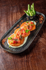 A gourmet dish of crispy potato pancakes topped with smoked salmon, garnished with fresh herbs and green onions, served on a sleek black plate with a side of cucumber salad