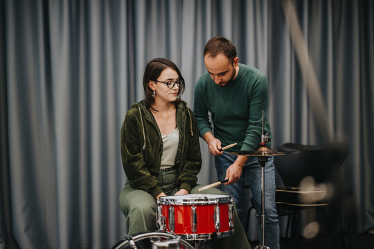 A male instructor attentively helps a female student practice drumming skills in a quiet rehearsal studio, emphasizing music education and collaboration to develop their percussion techniques.