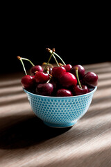 Bowl of ripe red cherries in a blue textured bowl, lit by natural light with strong shadows on a wooden table and a dark background