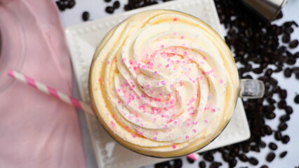 Pink dessert coffee topped with whipped cream and pink sugar crystals. Fresh hot coffee in a mug and a pink stripped straw surrounded by coffee beans.  Delicious and sweet coffee with cream and sugar