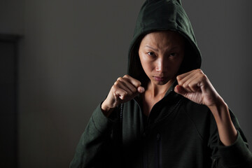 Mid adult Asian woman raising fists in dim training room wearing green hooded sweatshirt