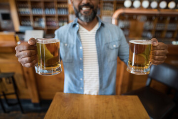 Mature Indian man wearing denim shirt holding two frothy beer mugs at wooden bar counter
