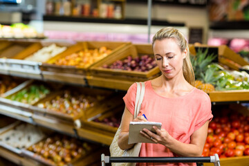 Woman writing shopping list on notepad in produce aisle with canvas tote bag, copy space