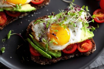 Delicious open-faced avocado toast topped with sunny side up egg and cherry tomatoes on a black plate