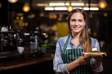 Woman wearing green apron presenting dessert plate with strawberry cream at coffee shop, copy space