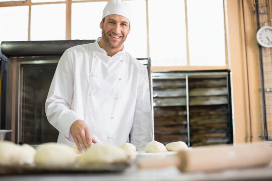 Male baker rolling dough balls with rolling pin on steel worktable by oven at bakery kitchen