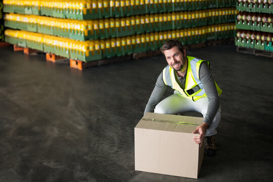 Male warehouse worker wearing vest kneeling in warehouse lifting box beside crates, copy space