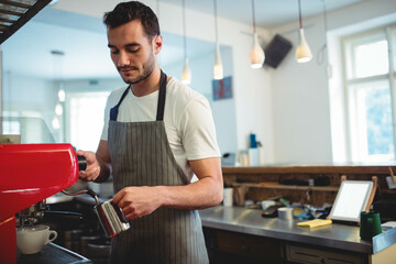 Male barista steaming milk with metal pitcher behind espresso machine at coffee shop counter