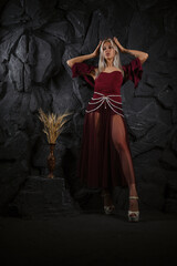 Beautiful woman posing a medieval queen in burgundy dress with gothic crown on her head posing on a dark black wall background...