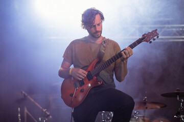Obraz premium Male musician playing electric guitar on raised stage near drum kit under blue spotlights with fog