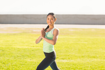 Asian woman standing on grassy lawn holding prayer pose against concrete wall under bright sunlight