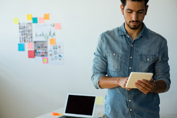 Middle Eastern pro standing reviewing tablet in modern office with sticky notes collage and laptop