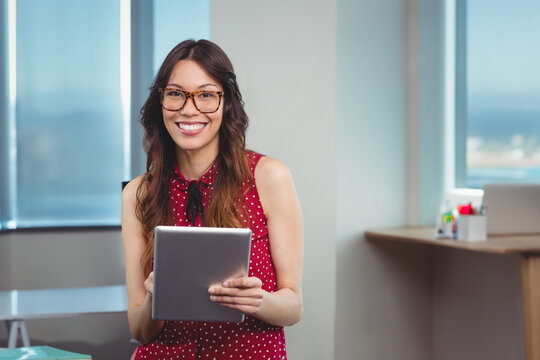 Adult woman wearing polka dot blouse and eyeglasses holding tablet at office desk by large windows - Powered by Adobe