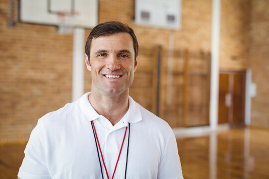 Male coach standing and smiling in school gymnasium wearing red-black lanyard and whistle