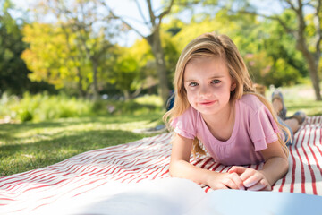 Child girl reading open book on red and white striped blanket at grassy park, copy space