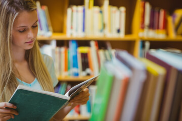 Teenage girl reading open book in library aisle with bookshelves and blurred books, copy space
