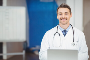 Male doctor sitting at clinic desk wearing stethoscope and using laptop near whiteboard