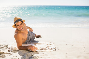 Man wearing straw sun hat, grey swim trunks reclining on beach towel at shoreline, copy space