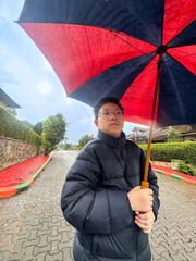 Asian boy holding a large red and blue umbrella on a wet residential street during rainy weather....