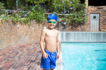 Boy standing at edge of outdoor pool on brick deck wearing blue swim cap and goggles