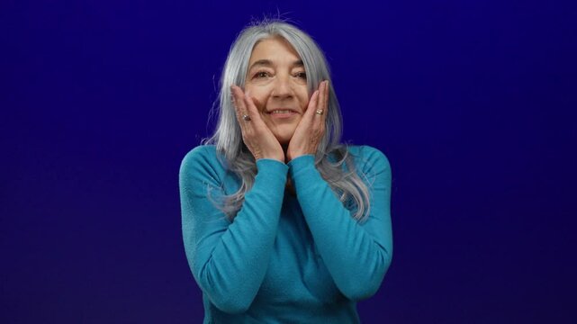 Woman with gray hair wearing blue sweater looks surprised against a vivid blue background, expressing surprise and curiosity.