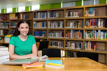 Woman studying at library table wearing green shirt with open book, laptop, notebooks, copy space