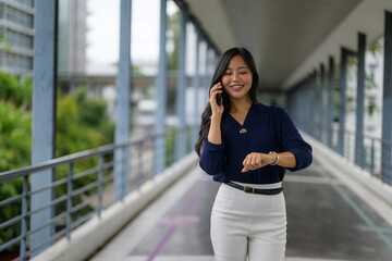 Fototapeta premium Young Asian businesswoman walking in a modern office building, talking on her phone and checking the time on her watch
