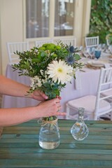 Florist woman preparing flower arrangements for a celebration, placing a bouquet of white and green flowers in a glass vase on a wooden table