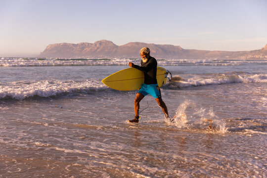 Senior African American man walking through water at beach carrying surfboard in board shorts