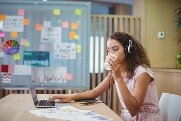 Woman wearing light pink dress and headphones, sipping coffee, reviewing charts on laptop at office