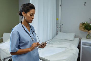 Mature nurse wearing blue scrubs and stethoscope standing beside bed in patient room holding tablet