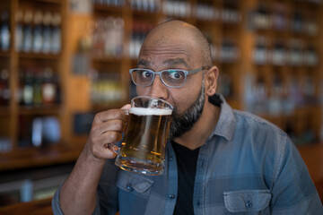 Indian man wearing eyeglasses lifting clear glass mug with beer foam while sitting at pub counter