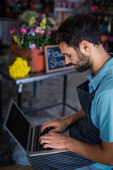 Bearded florist typing on laptop at flower shop workspace with wooden crates of colorful blooms