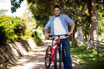 Obraz premium Man wearing helmet pushing red bicycle along sunlit park path with stone wall and park bench