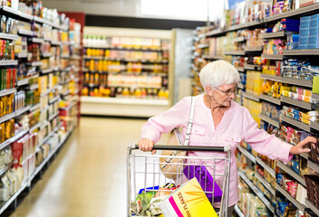 Senior woman pushing shopping cart down supermarket aisle picking cereal box from shelf