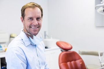 White male dentist standing smiling with mask around neck in treatment room beside red dental chair