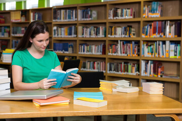 Female student in green shirt reading at library with laptop and books, copy space