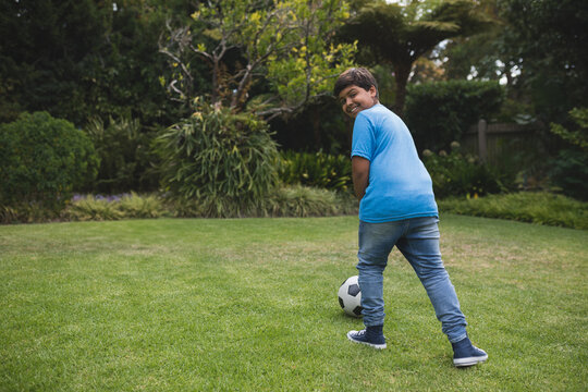 Indian boy looking over shoulder on backyard lawn while resting foot on soccer ball, copy space