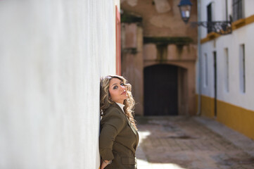 Fototapeta premium Blonde woman with curly hair, young and beautiful, rests her back and hands on a white wall. The woman is happy and serene.